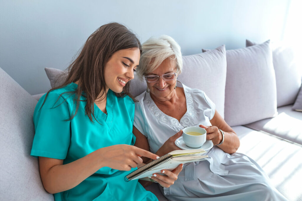 Dominion of Louisville | Senior woman stinng on a couch with her caretaking while holding a cup of tea and looking at some notes